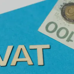 Drewniane litery VAT na niebieskim tle, obok banknot 100 zł i monety. Podatek VAT to ważny element finansów.