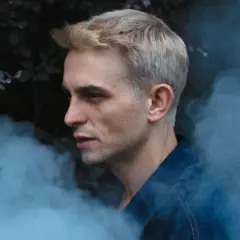 Vito Bambino w dymie, zapowiada nową płytę. Artysta w denimowej kurtce, z blond włosami, na tle ciemnych liści.