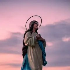 Figurka Matki Boskiej z aureolą na tle różowego nieba. W dłoniach trzyma błękitną szatę, jakby w modlitwie "Anioł Pański".