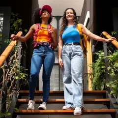 Dos amigas con estilo y2k, una con top naranja y gorra rosa, la otra con top azul, bajan unas escaleras rodeadas de plantas.