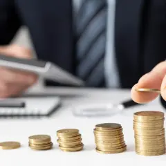 Dłoń układa monety, budując stosy. W tle mężczyzna z kalkulatorem, sugerującym planowanie finansowe i poszukiwanie optymalnych rozwiązań, być może związanych z **rajami podatkowymi**.