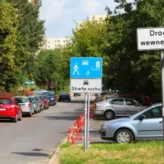 Droga wewnętrzna z oznaczeniem strefa zamieszkania. Samochody parkują wzdłuż ulicy, otoczonej drzewami.