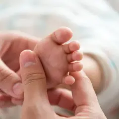 Dłoń delikatnie bada stopę niemowlęcia, wywołując objaw Babińskiego.