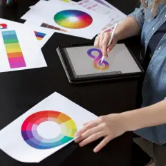 Grafik tworzy projekt, korzystając z próbek kolorów i tabletu. Na stole leżą wydruki z kołami barw i paskami kolorów, a na ekranie tabletu powstaje logo.