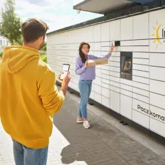 Mężczyzna z telefonem w ręku obserwuje kobietę, która wkłada paczkę do Paczkomatu InPost. Czy paczkomat InPost drukuje etykiety? Nie, ale ułatwia nadawanie.