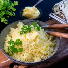 En skål med fluffig pressad potatis, toppad med en smörklutt och färsk persilja. Bredvid syns en smörskål och en rivare.
