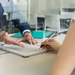 Podpisanie dokumentów w urzędzie. Dłoń z długopisem nad umową, obok płyn do dezynfekcji. Pracownik budżetówki wskazuje na treść.