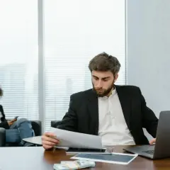 A man in a suit reviews documents, perhaps researching accelerated online MPA programs. A woman in the background is on her phone.