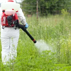 Osoba w białym kombinezonie używa opryskiwacza plecakowego spalinowego do zwalczania chwastów na łące.
