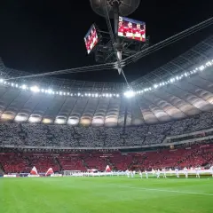 Stadion Narodowy w Warszawie, z imponującą pojemnością, wypełniony kibicami w biało-czerwonych barwach. Na telebimach widać piłkarzy.
