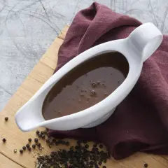 En vit såsslev fylld med mörk brunsås, bredvid en hög med svartpepparkorn på en träskärbräda. Perfekt för ditt brunsås recept.