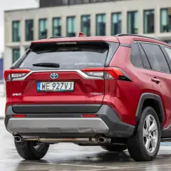 Czerwony, używany Toyota RAV4 Hybrid z salonu, zaparkowany na mokrym parkingu przed nowoczesnym budynkiem.