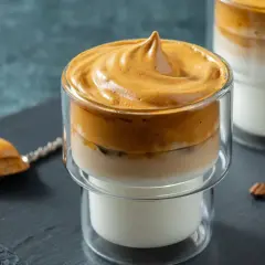 Puszyste dalgona coffee w szklance, z pianką na wierzchu i mlekiem na dole. Obok łyżeczka z pianką i ziarna kawy.