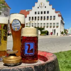 Drei Gläser Bier, darunter ein helles und ein dunkles, stehen auf einem Fass. Im Hintergrund eine malerische Stadtkulisse mit einem historischen Gebäude. Ein perfekter Genuss von italienischem Bier.