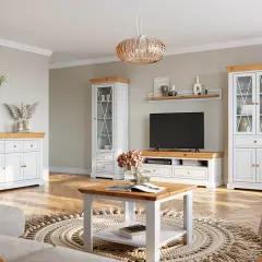 Przytulny mały salon w bloku ze stołem kawowym. Białe meble z drewnianymi blatami, wiszące rośliny i okrągły dywan tworzą ciepłą atmosferę.