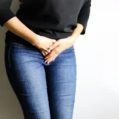 Femme en jean tenant ses mains sur son bas-ventre, évoquant une gêne, peut-être liée à une bronchite et une fuite urinaire.