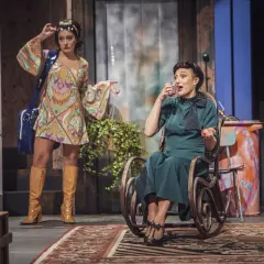 Dwie kobiety na scenie: jedna w kolorowej sukience i kozakach, druga siedzi w fotelu, pijąc drinka.