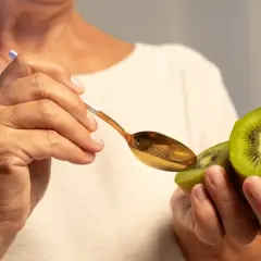 Dłonie trzymają połówkę kiwi i złotą łyżeczkę. Owoc jest bogaty w witaminy, np. C i K.
