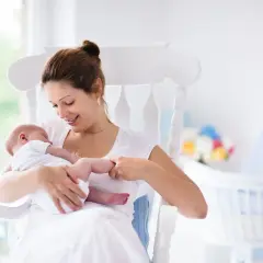 Uśmiechnięta mama kołysze noworodka. W tle widać łóżeczko i kwiaty. To może być moment, gdy zastanawiasz się, jak zatrzymać laktację.