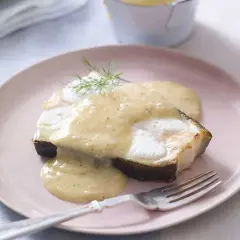 En bit fisk täckt med krämig beurre blanc-sås, garnerad med dill, serveras på en rosa tallrik med en gaffel.