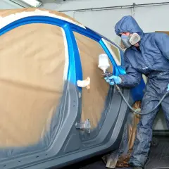 Pittore in tuta blu spruzza vernice su un'auto blu. Il costo per cambiare il colore della macchina sul libretto è un dettaglio importante.