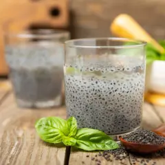 Due bicchieri con semi di basilico in acqua, un cucchiaino di semi e foglie fresche su un tavolo di legno.