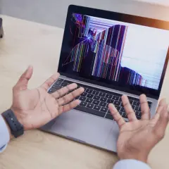 Rozlany ekran w laptopie wyświetla kolorowe pęknięcia, a osoba z rozłożonymi rękami patrzy na niego z niedowierzaniem.