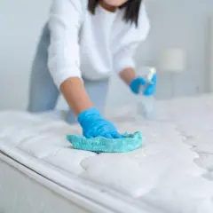Person mit blauen Handschuhen reinigt die Matratze mit einem Tuch und Sprühflasche.