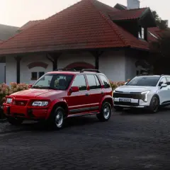 Czerwony, klasyczny Kia Sportage 1 obok białego, nowoczesnego SUV-a. Dwa pokolenia na tle urokliwego budynku.