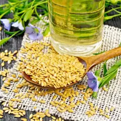 Semi di lino dorati su un cucchiaio di legno, con una bottiglia di olio di lino e fiori di lino sullo sfondo.