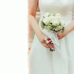 Panna młoda trzyma bukiet z białych róż i gipsówki, ozdobiony białą wstążką. Klasyczne rodzaje bukietów ślubnych.