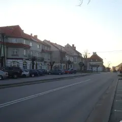 Ulica w miasteczku, gdzie są Wydminy. Rząd kamienic z czerwonymi dachami, zaparkowane samochody i droga.