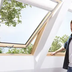 Mężczyzna otwiera okienko w dachu, przez które widać zielone drzewa i niebo.