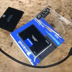 Dwa dyski SSD, jeden Samsung, drugi Goodram CX400 256 GB, obok obudowa na dysk i kable SATA.