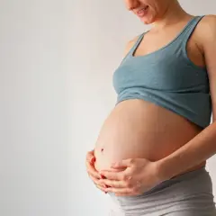 Uśmiechnięta ciężarna kobieta trzyma dłonie na brzuchu. Wkrótce może pojawić się czop śluzowy.