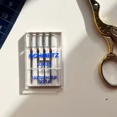 Schmetz Jersey needles, size 90/14, are ideal for stretch fabrics. A blue measuring tape and gold scissors complete the sewing setup.