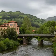 Un puente cruza un río, conectando pueblos bonitos cerca de Liérganes. Edificios rosados y verdes colinas completan la escena.