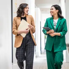 Zwei Frauen in lässiger Business-Kleidung gehen durch einen Flur. Eine trägt einen braunen Blazer und karierte Hose, die andere einen grünen Hosenanzug.