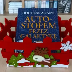 Książka "Autostopem przez galaktykę" Douglasa Adamsa w czerwznym, świątecznym koszyku z reniferem i choinkami.
