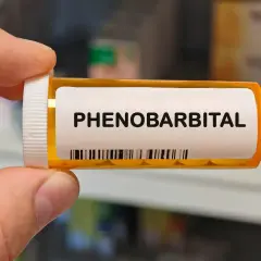 Dłoń trzyma buteleczkę z lekiem. Na etykiecie widnieje napis "PHENOBARBITAL" i kod kreskowy.