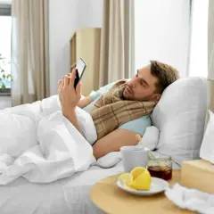 Chory mężczyzna w łóżku, owinięty szalikiem, przegląda telefon. Obok kubek z herbatą, cytryna i miód. Czyżby czekał na zwolnienie lekarskie z powodu siły wyższej, zgodnie z kodeksem pracy?