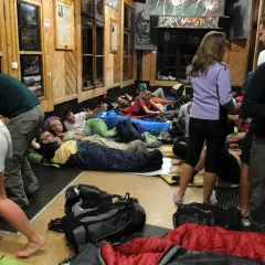 Ludzie śpią na materacach w schronisku górskim, otoczeni plecakami i śpiworami.