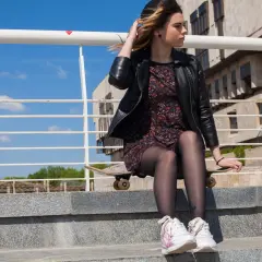 Dziewczyna w eleganckiej sukience i sportowych butach siedzi na deskorolce na schodach.