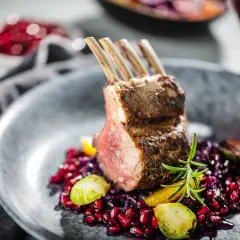 Carré d'agneau parfaitement rôti, servi avec une garniture colorée de grenade, chou rouge et romarin. La cuisson du carré d'agneau est mise en valeur par la présentation.