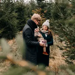 Rodzina podczas sesji świątecznej w plenerze, wśród choinek. Tata trzyma dziecko, mama uśmiecha się, trzymając ozdobne pudełko.