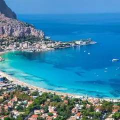 Vista aerea della spiaggia di Palermo, con acque turchesi, case colorate e montagne sullo sfondo.