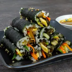 Gestapeltes Kimbap, koreanische Reisrollen mit Gemüse und Ei, auf schwarzer Platte, daneben eine Schale Dip.