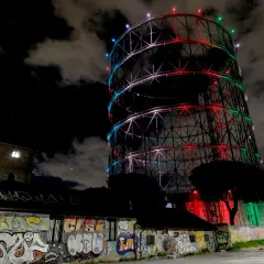 Il gasometro di Roma, illuminato da luci colorate, si staglia contro il cielo notturno. Un muro di graffiti decora l'edificio sottostante.