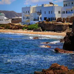 Blågrönt hav sköljer mot klippor och sandstrand. Vita hus med gröna tak reser sig mot en bergig bakgrund på Naxos, Grekland.