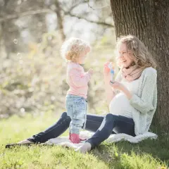 Ciężarna kobieta i mała dziewczynka puszczają bańki pod drzewem. Nadopiekuńcza matka patrzy z uśmiechem na swoje dziecko.